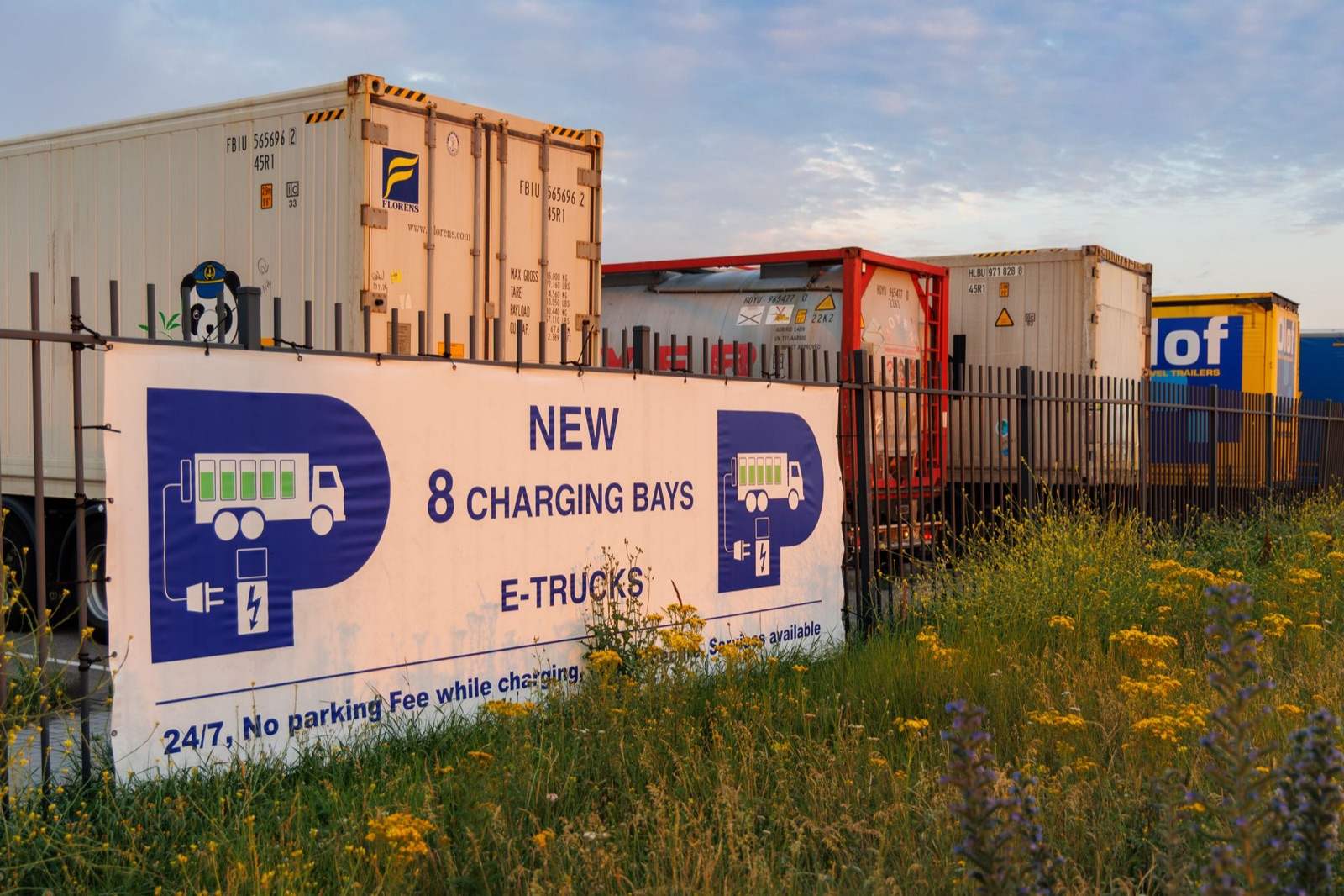 Bord bij een truckparking in Rotterdam met acht laadplekken voor elektrische vrachtwagens, met containers erachter